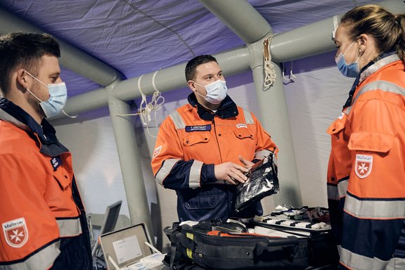 Eine Frau und zwei Männer in der Bekleidung der Malteser und mit Mundschutz und prüfen Medikamententaschen bzw. -koffer.