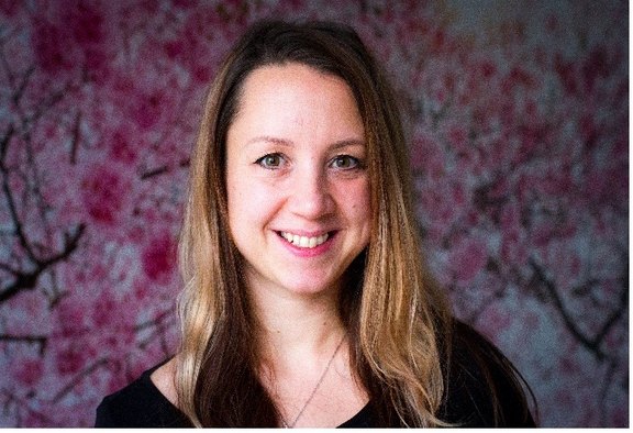 Das Foto zeigt den Kopf einer jüngeren Frau, die in die Kamera lächelt. Sie hat braune längere Haare. Das Oberteil, das sie trägt ist schwarz. Im Hintergrund ist rosa blühende Zweige zu sehen.