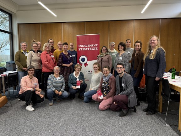 Das Foto zeigt 20 Frauen und Männer , die sich in einem Seminarraum um das Rollup der Engagementstrategie gruppiert haben und in die Kamera lächeln. Es sind die Teilnehmenden an einer Fortbildungsveranstaltung.