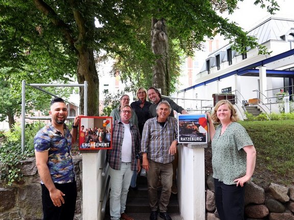 Vertreter*innen aus Ratzeburg strahlen gemeinsam mit Catarina Mierwald in die Kamera. Sie stehen unter Bäumen auf einen kurzen Treppe.