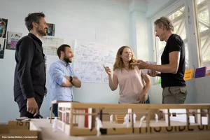 drei jüngere Männer und eine Frau stehen zwischen einem Tisch und einer Wand mit einem Mindmap auf einem großen Papier und diskutieren miteinander.