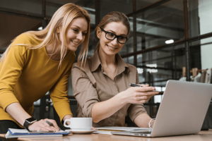 Zwei jüngere Frauen schauen gut gelaunt auf ein aufgeklapptes Laptop.