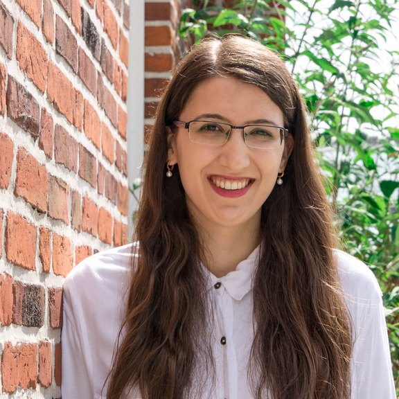 Das Foto zeigt eine junge Frau mit Brise und langen braunen Haaren, die eine helle Bluse trägt, an einer roten Backsteinmauer lehnt und in die Kamera lächelt.