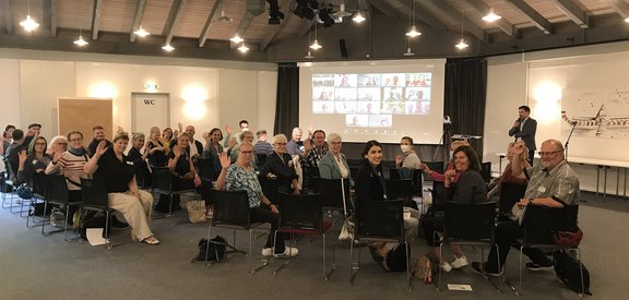 Das Foto zeigt gut 20 Veranstaltungsteilnehmende, die sich auf ihren Stühlen umdrehen und in die Kamera winken. Vorne auf der Leinwand sind per Internet zugeschaltete Teilnehmende zu sehen. Ein Referent steht rechts neben der Leinwand.