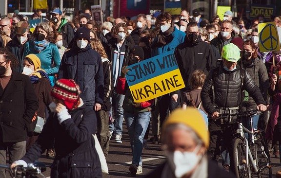 Das Foto zeigt Teile eines Demonstrationszuges. Ein Schild ist zu sehen mit den Farben der Ukraine und den Worten: Stand with Ukraine.