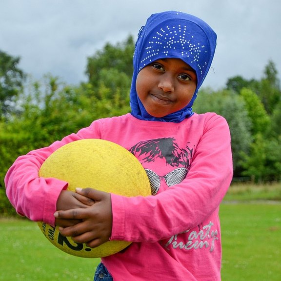 Das Foto zeigt ein kleines Mädchen mit dunkler Hautfarbe, es trägt eine blaue muslimische Kopfbedeckung, lächelt in die Kamera und trägt im rechten Arm einen gelben Ball.
