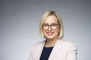 Das Foto zeigt die Staatsministerin Dr. Schenderlein. Sie trägt ein dunkles Oberteil mit weißem Jacket und eine dunkle Brille. Sie lächelt in die Kamera.