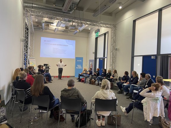Auf diesem Foto sitzen ca. 30 Teilnehmende an einer Veranstaltung in einem schmalen Raum in einem offenen Oval, der Referent steht in weißen Hemd und dunkler Hose vor einer Leinwand, auf der eine Präsentation zu sehen ist. Recht ist eine Rollup und links eine beschriebene Plipchart zu sehen.