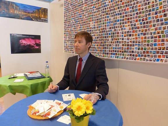 In dem Bild steht ein Mann mit Jacket und Schlips in einem Messestand vor einer bunten Wand an einem Stehtisch mit blauer Decke, Infomaterial und gelben Blumen.