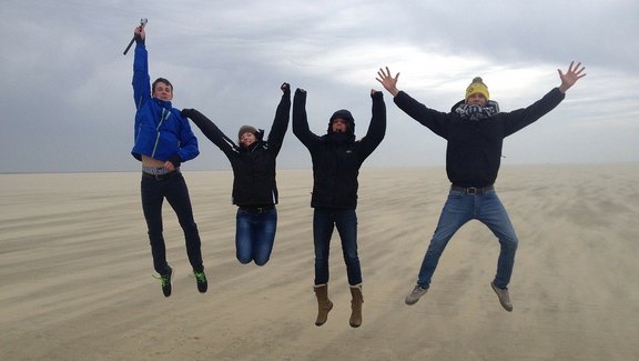 Zu sehen sind in dem Bild vier junge Leute an einem herbstlichen Strand, die sich offensichtlich freuen und gleichzeitig dabei in die Luft springen.