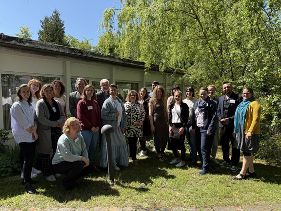 Foto: Eine Gruppe von 22 Frauen und Männern stehen auf einer kleinen Rasenfläche vor einem niedrigen Gebäude und grünen Büschen und Bäumen. Alle lächeln in die Kamera.