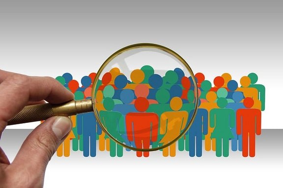 Ein Hand hält ein Vergrößerungsglas und betrachtet eine bunte Gruppe von stilisiert dargestellten Personen.