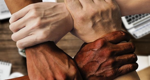 Das Foto zeigt vier Hande, die sich jeweils am Handgelenk festhalten und so ein geschlossenes Rechteck bilden. Zu sehen ist, dass es Hände von Menschen verschiedener Hautfarbe sind.