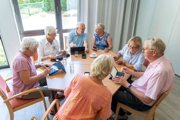 Sieben Senior*innen sitzen mit ihren iPads an einem runden Tisch und sprechen mit einander