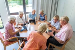 Sieben Senior*innen sitzen mit ihren iPads an einem runden Tisch und sprechen mit einander 