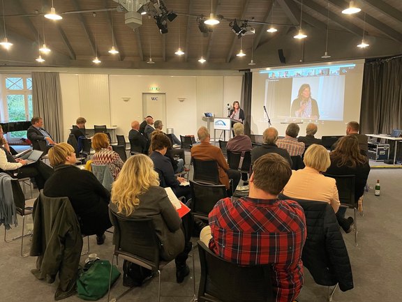 Foto: 20 Teilnehmende sitzen auf Stühlen und schauen nach vorne zu einer Referentin am Rednerpunkt, die zugleich auf der Leinwand dahinter auch zu sehen ist.