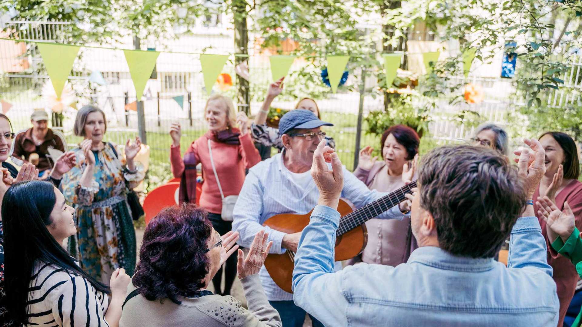 Zu sehen ist eine bunte Gruppe von Leuten, die miteinander Spaß haben; in der Mitte steht ein Mann mit einer Gitarre und spielt.