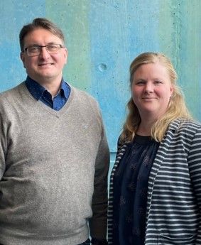 Das Foto zeigt links einen Mann mit Brille und grauem Pulli und blauem Hemd, rechts eine blonde Frau mit schwarzem Top und schwarzgrau quergestreifter leichter Jacke. Beide lächeln in die Kamera.