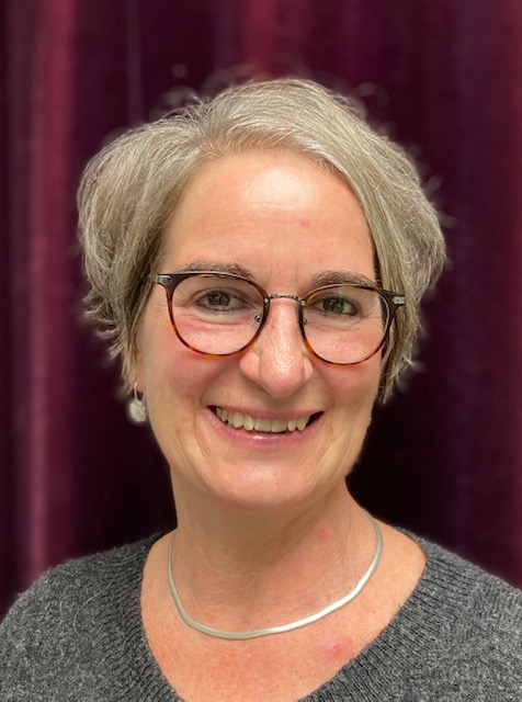 Das Foto zeigt eine in die Kamera lächelnde Frau. Sie hat graue gescheitelte kürzere Haare, eine rote Brille und einen Ohrschmuck mit einer Perle. Ihr Halsschmuck liegt eng an und besteht aus grauem Metall.