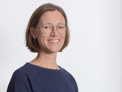 Das Foto zeigt den Oberkörper einer jüngeren Frau, leicht nach rechtsgedreht, der Kopf zur Kamera gewendet. Sie trägt eine Brille mit dünnem Rand, ihre braunen Haare fallen kinnlang, das Oberteil ist dunkelblau.