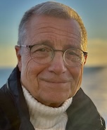 Das Foto zeigt einen etwas älteren Mann, angegraute kurze Haare, Brille. Er einen weißen Rollkragenpullover, eine dunkelblaue Windjacke und lächelt freundlich in die Kamera. Im Hintergrund ist blauer Abendhimmel und das Kielwasser eines Schiffes zu erkennen.